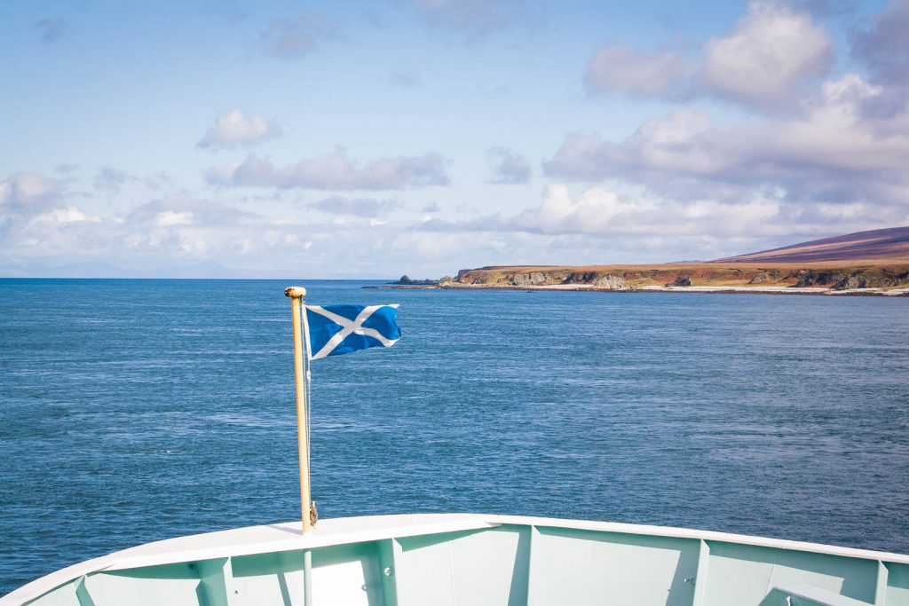 Scotland flag at the front of a ferry with and island in the background.
