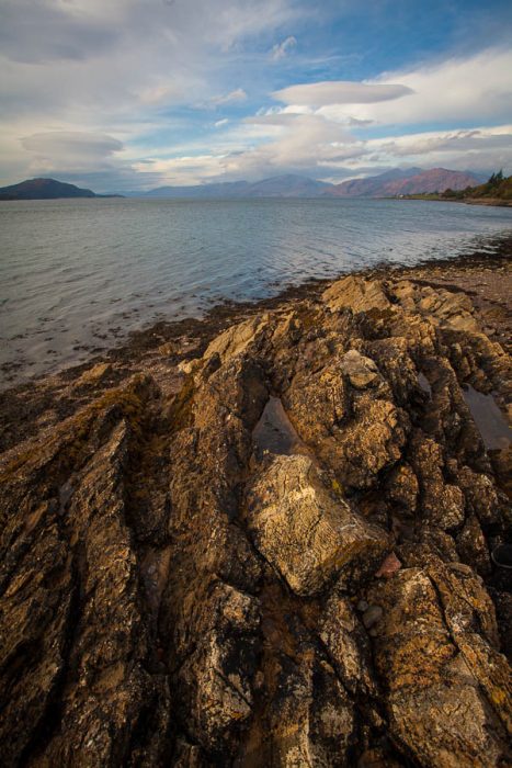 A rocky beach by a loch and mountains in the background.