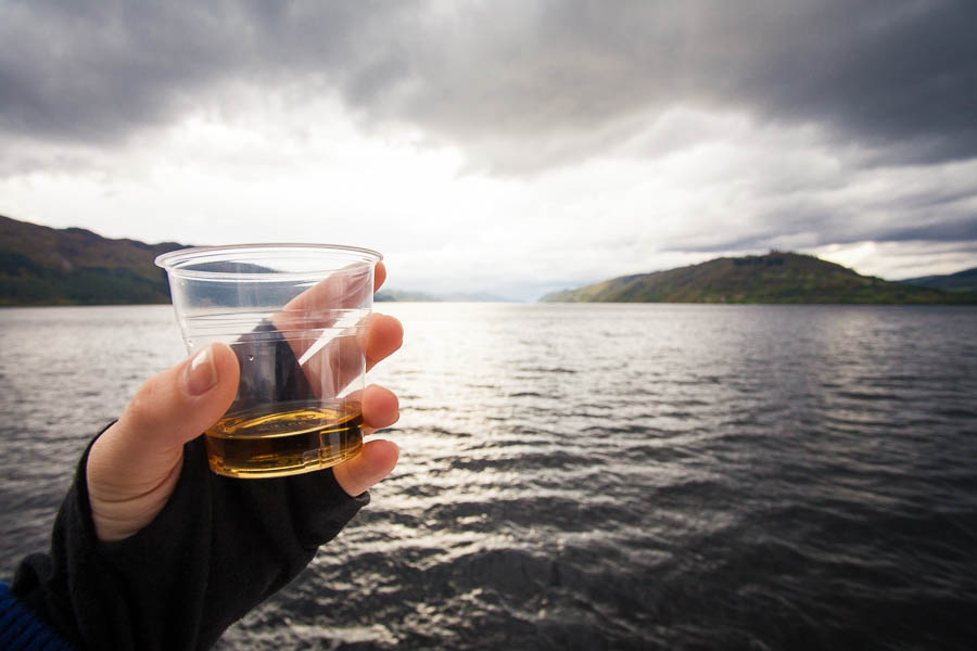 A hand holding a glass of whisky and Loch Ness in the background.