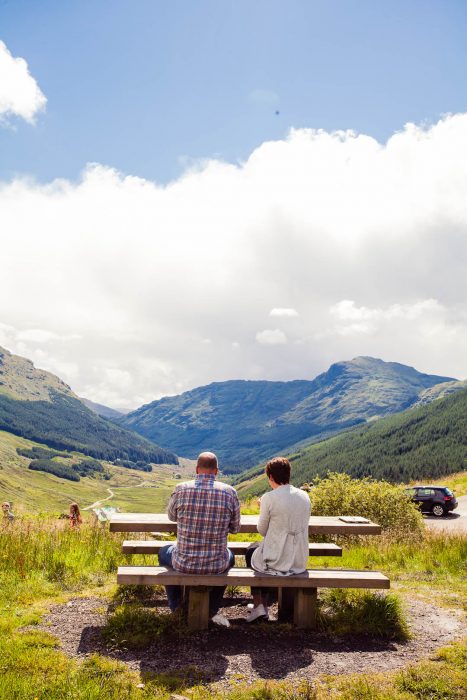 A couple sitting on a bench in the Scottish Highlands overlooking the mountains and valley.