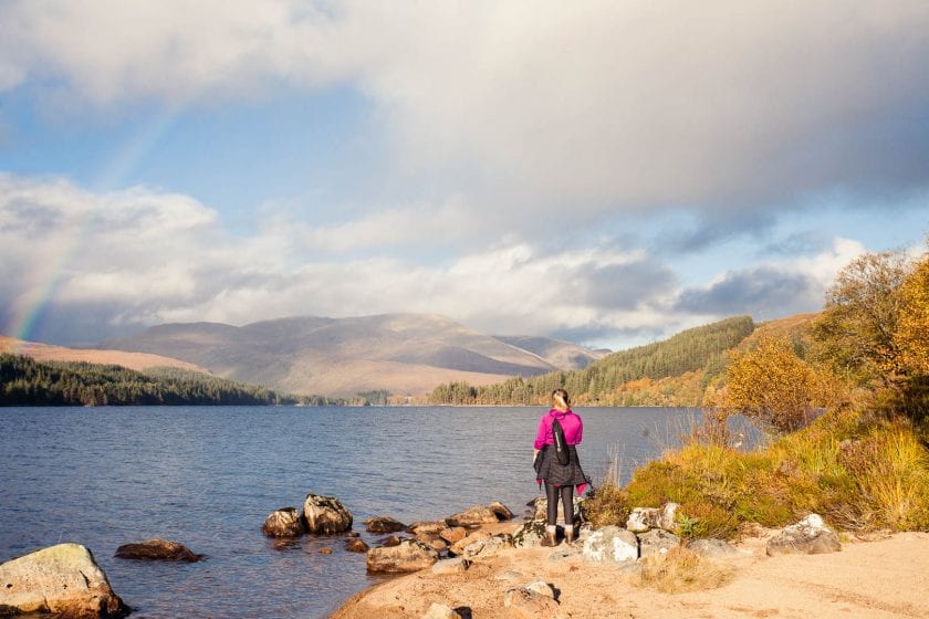 Woman standing by a lake and rainbow at Loch Ossian Youth Hostel