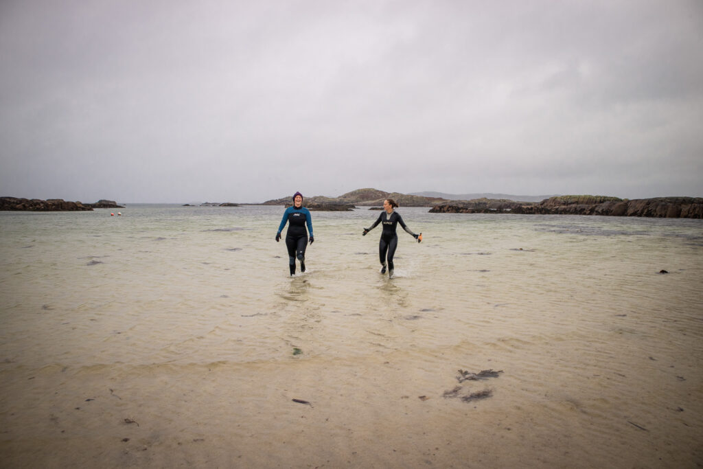 two women in wet suits walking out of the ocean in scotland